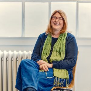 Karis, a white woman with short blond hair and sunglasses on her head, stands in front of house in the sun, smiling.