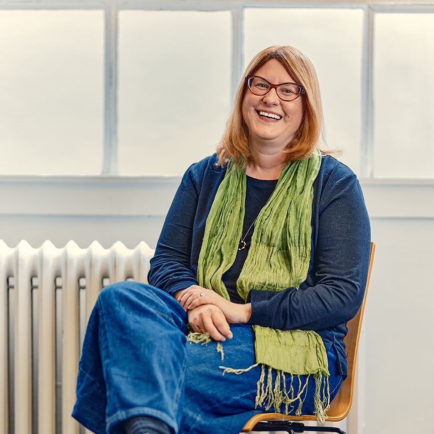 Karis, a white woman with short blond hair and sunglasses on her head, stands in front of house in the sun, smiling.