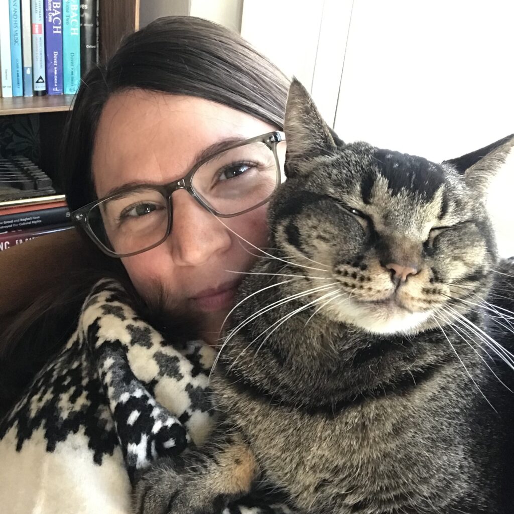 Jill, a white woman with brown hair and glasses wearing a cozy sweater, cuddles her brown and black striped cat.