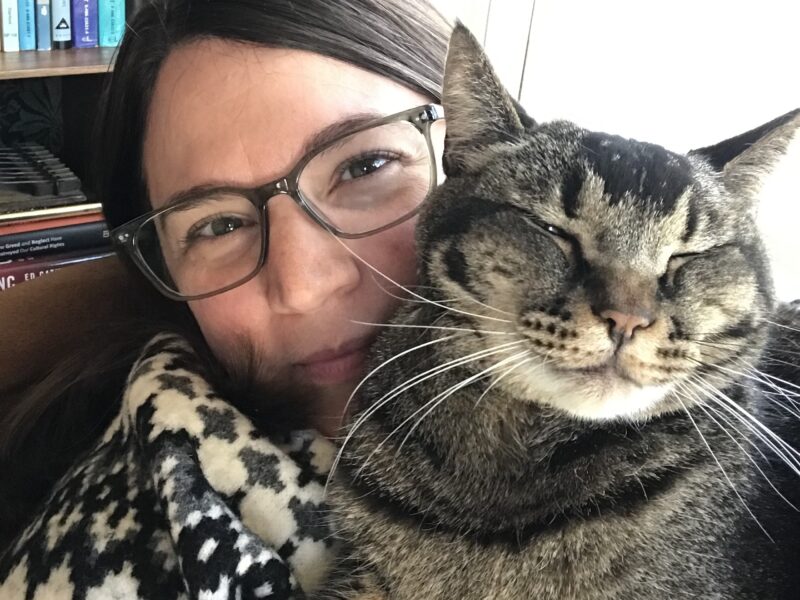 Jill, a white woman with brown hair and glasses wearing a cozy sweater, cuddles her brown and black striped cat.