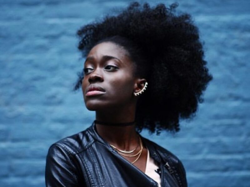 Holly, a black woman with an afro wearing an ear cuff and leather jacket, stands in front of a blue brick wall and looks away from the camera.