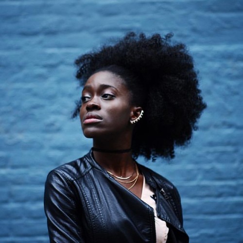 Holly, a black woman with an afro wearing an ear cuff and leather jacket, stands in front of a blue brick wall and looks away from the camera.