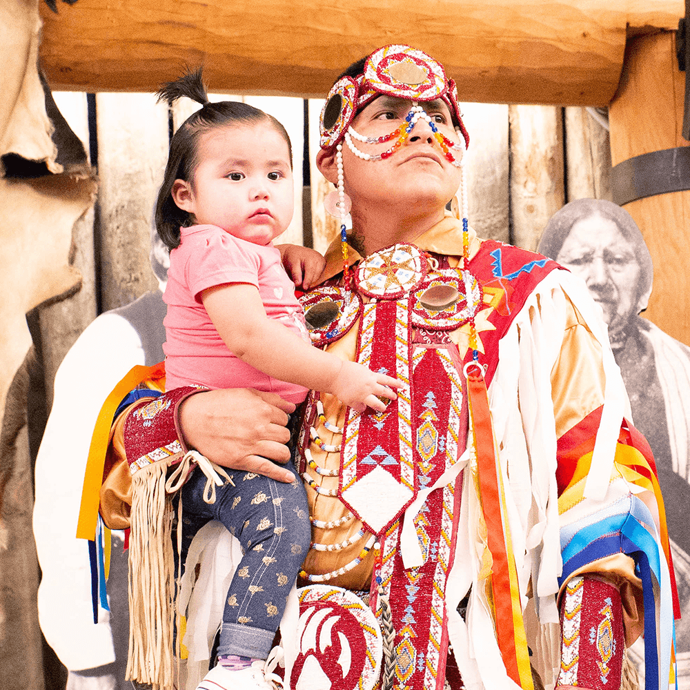 A Native person in full regalia looks beyond the camera. On his hip is a young Native child in jeans and a pink t-shirt.