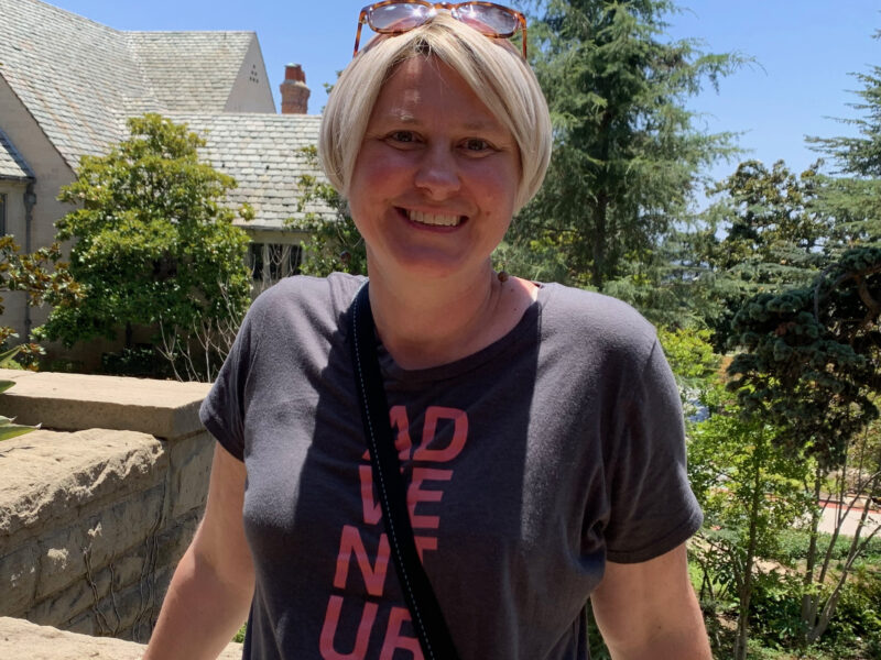 Karis, a white woman with short blond hair and sunglasses on her head, stands in front of house in the sun, smiling.