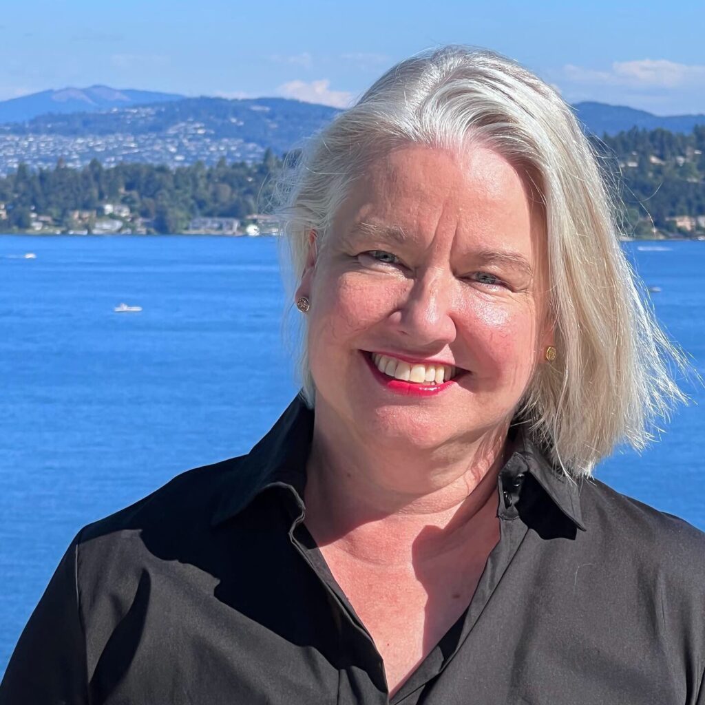 Barbara, a white woman with white hair and bright, pink lipstick, stands in front of the Puget Sound smiling.