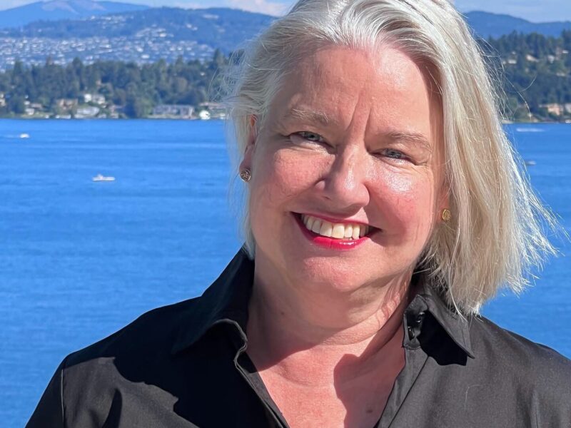Barbara, a white woman with white hair and bright, pink lipstick, stands in front of the Puget Sound smiling.