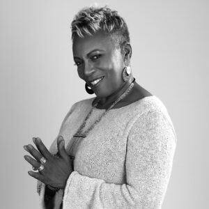 A black and white photo of Vivian, a Black woman with short hair and statement jewelry, looks at the camera and smiles.