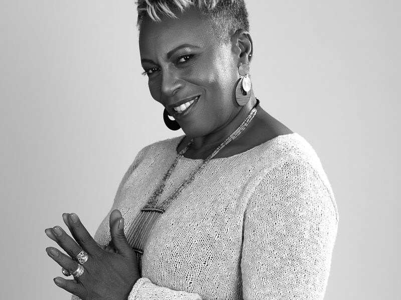 A black and white photo of Vivian, a Black woman with short hair and statement jewelry, looks at the camera and smiles.