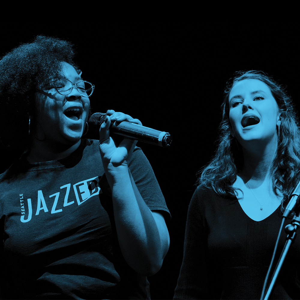 One black woman wearing a JazzEd t-shirt and one white woman in a black shit sing into microphones. The photo has a blue overlay.