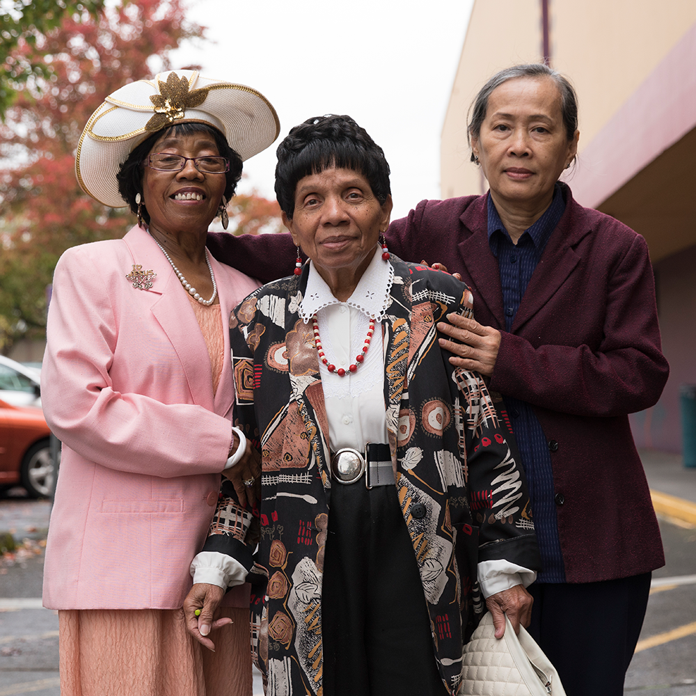 Two older black women and one older Asian women lean on each other with their arms around each other.