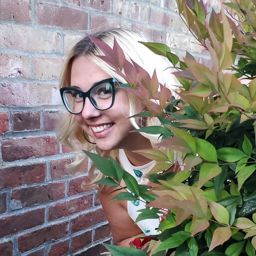 Image of Kira Shea, a white woman with blond hair and glasses, smiling and peeking out from behind a plant.