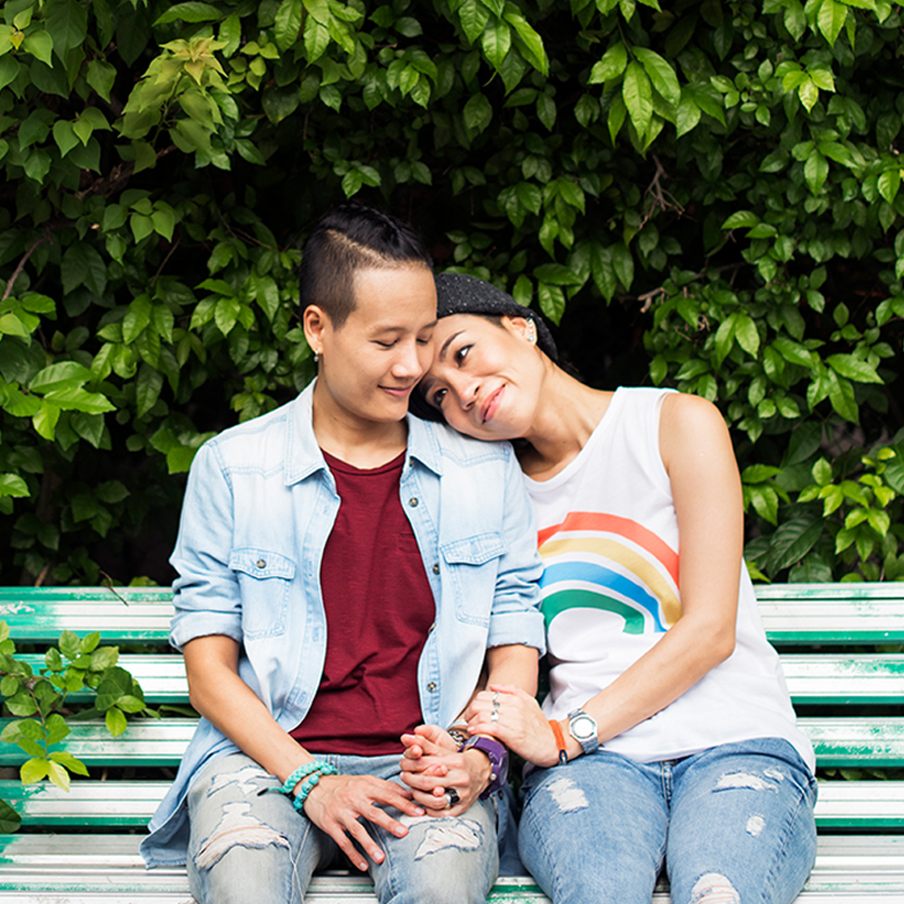 Two queer women of color sit on a bench, holding hands. One, who is wearing tank top with a rainbow on it leans against the other, who is wearing a red t-shirt and denim overshirt.
