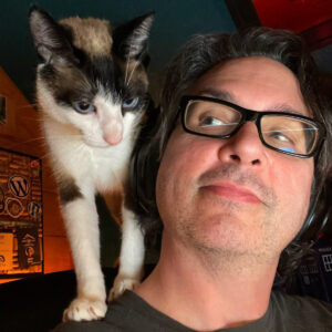 Jeff Brock, a white man with Brown hair and glasses wearing headphones, looks lovingly at his cat, Pi, who is white, brown, and black and is climbing on his shoulder.